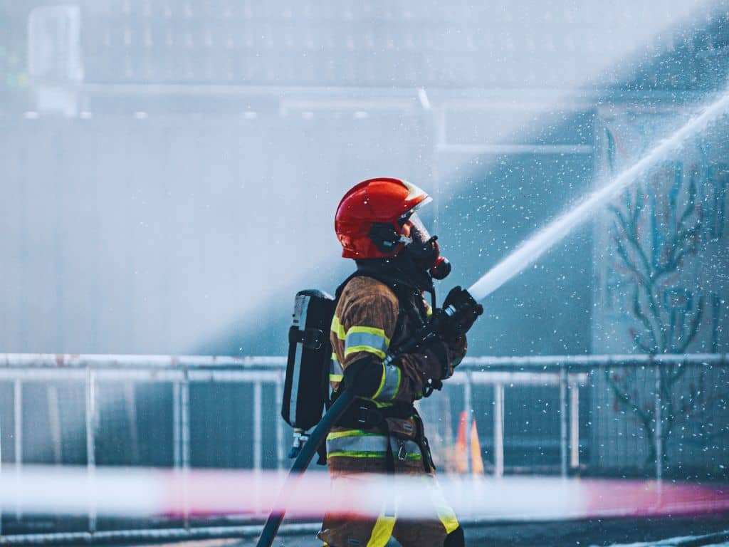 Firefighter in full gear spraying a water hose. The firefighter is in the middle of a hazy scene.